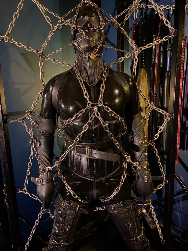 Encased in a latex catsuit and hood Mistress Servalan's slave is restrained to the chain web with leather and metal restraints.